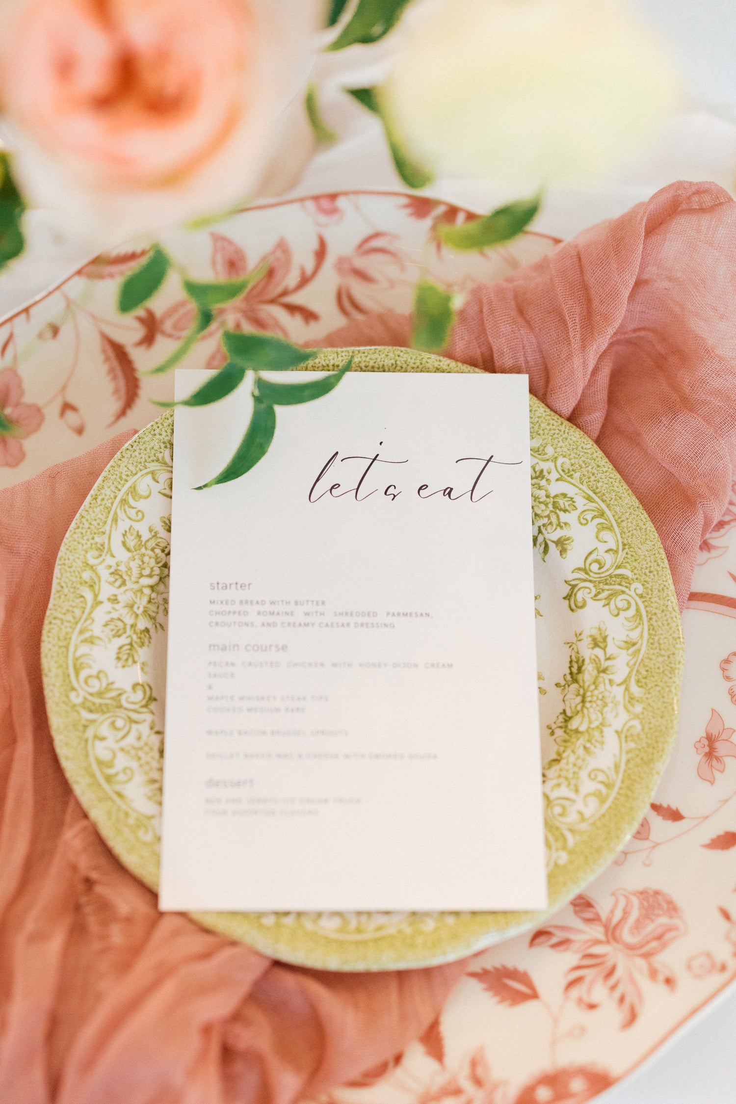 Decorative table setting with a floral plate and 'let's eat' card on a pink fabric background.