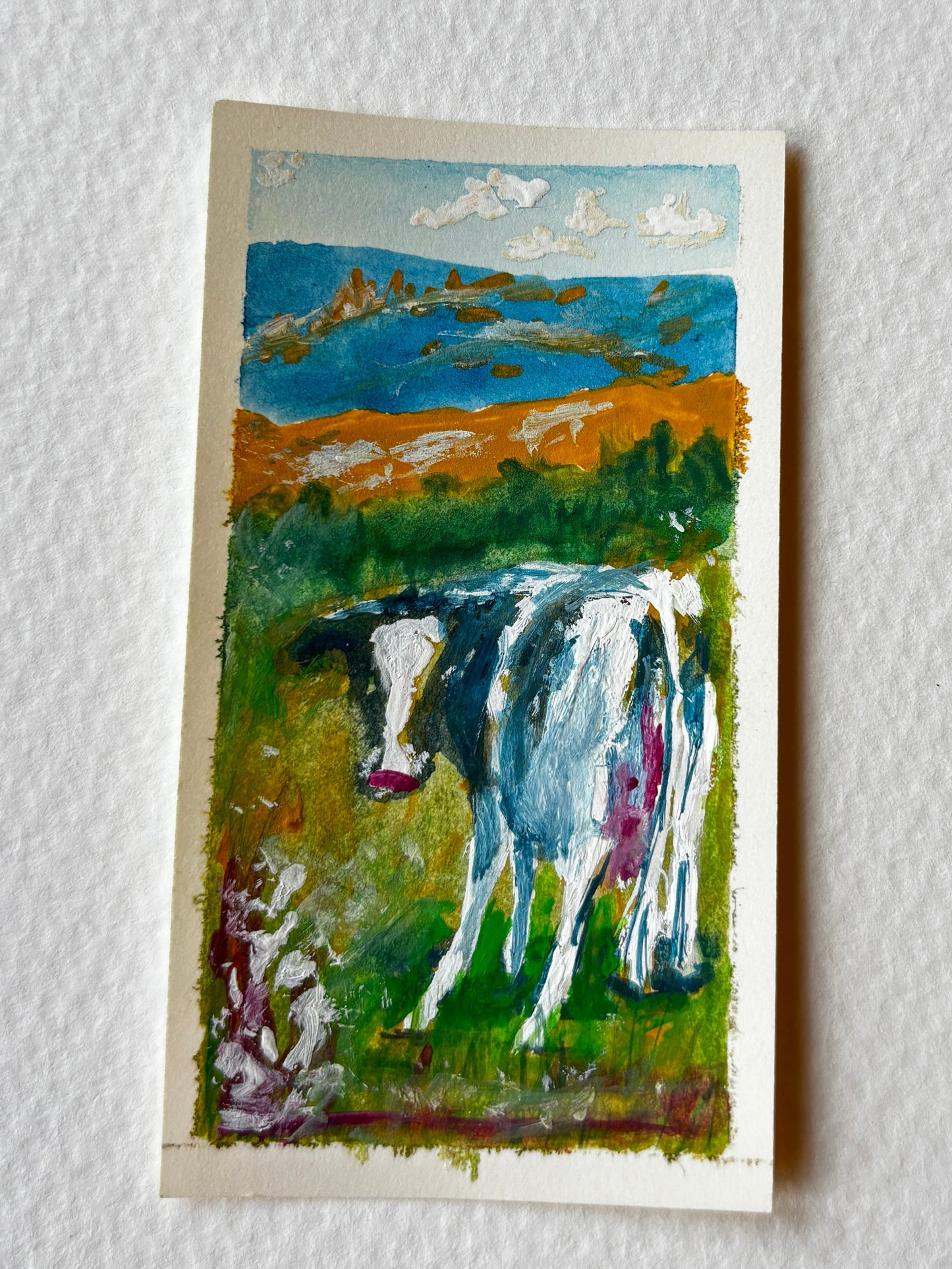 A small vertical painting of a cow in a grassy field with blue mountains and clouds, mounted on a textured white wall.