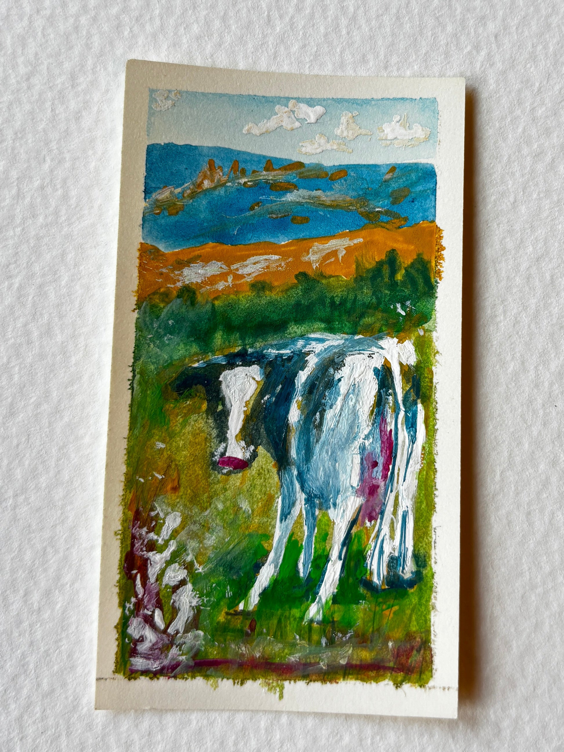 A small vertical painting of a cow in a grassy field with blue mountains and clouds, mounted on a textured white wall.