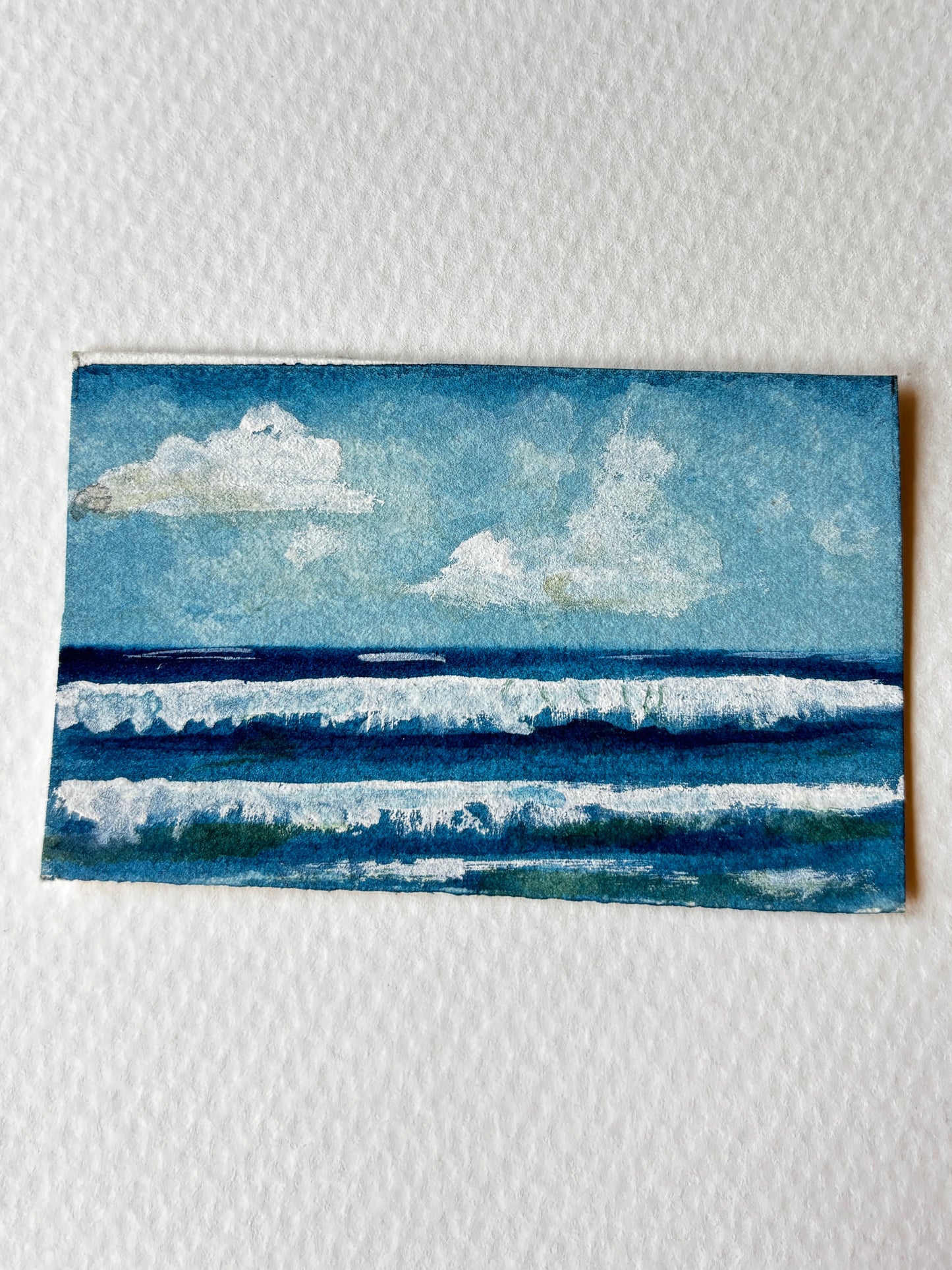 A small square watercolor painting of an ocean scene with blue waves and white clouds, placed on a textured white paper background.