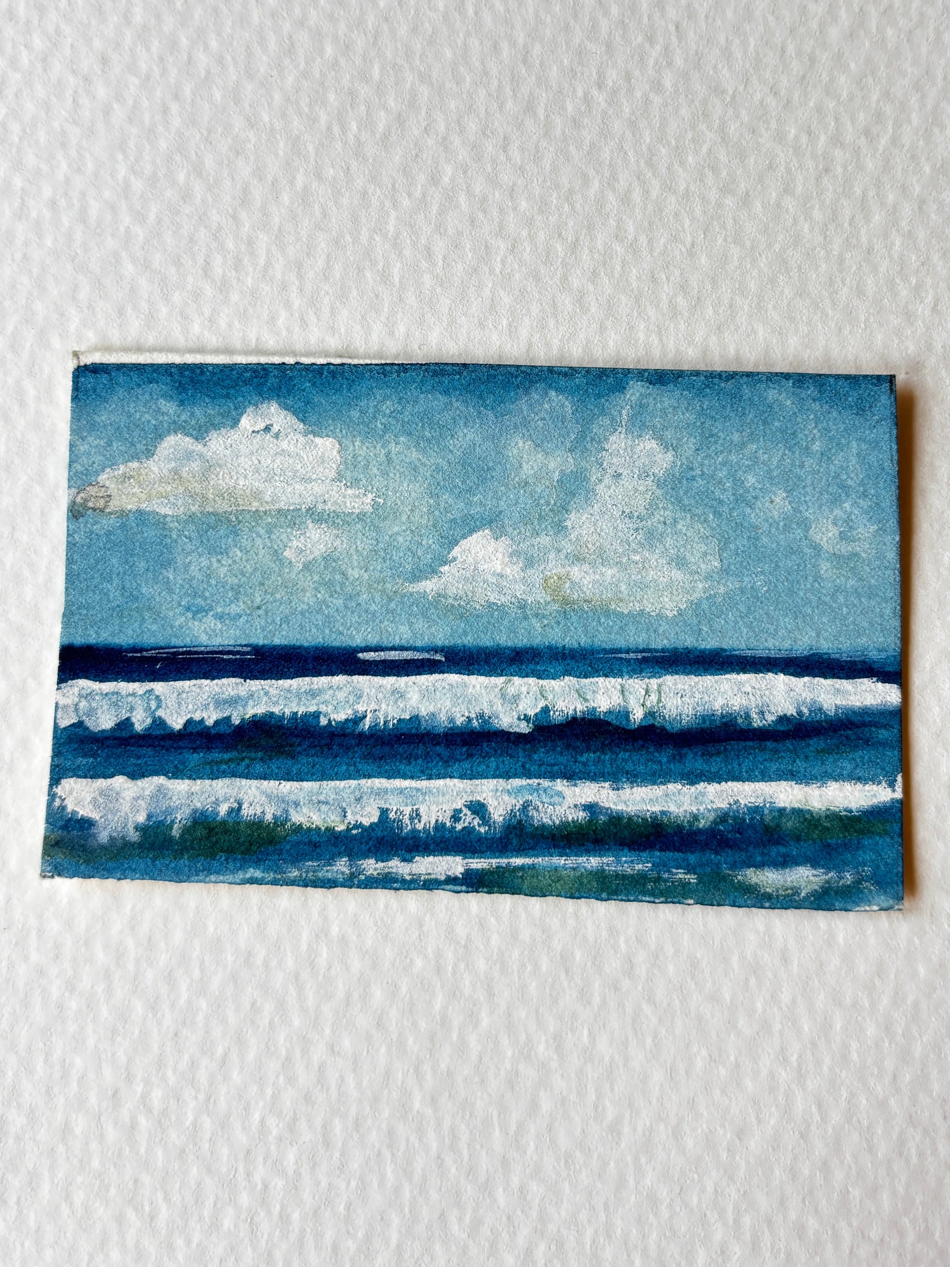 A small square watercolor painting of an ocean scene with blue waves and white clouds, placed on a textured white paper background.