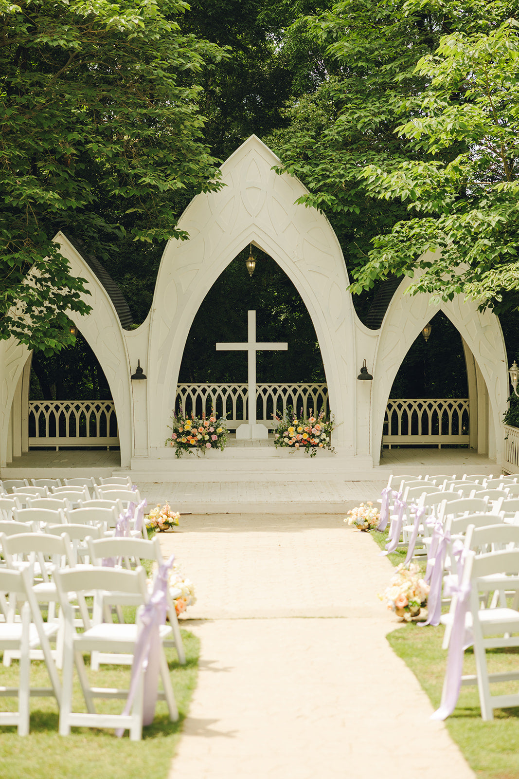 Outdoor wedding venue with white chairs, floral arrangements, and a cross on a white archway.